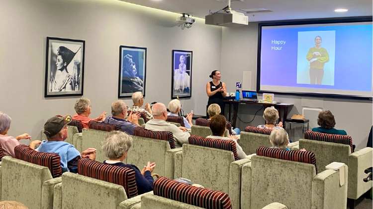 Residents listening to a community hearing awareness presentation in a lecture room, with a speaker and projected slide at the front.